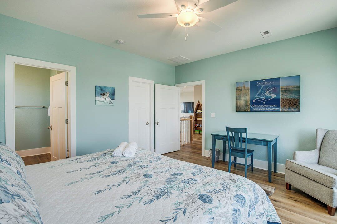A bedroom with a bed, a desk, and a chair. There is a ceiling fan and a door leading to another room.
