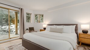 A modern bedroom with a large bed, two bedside tables, a nightstand, and a view of a snowy landscape through large windows. - ❄︎The Altitude❄︎ | 4 BR Renovated Luxury Townhome (Whistler)