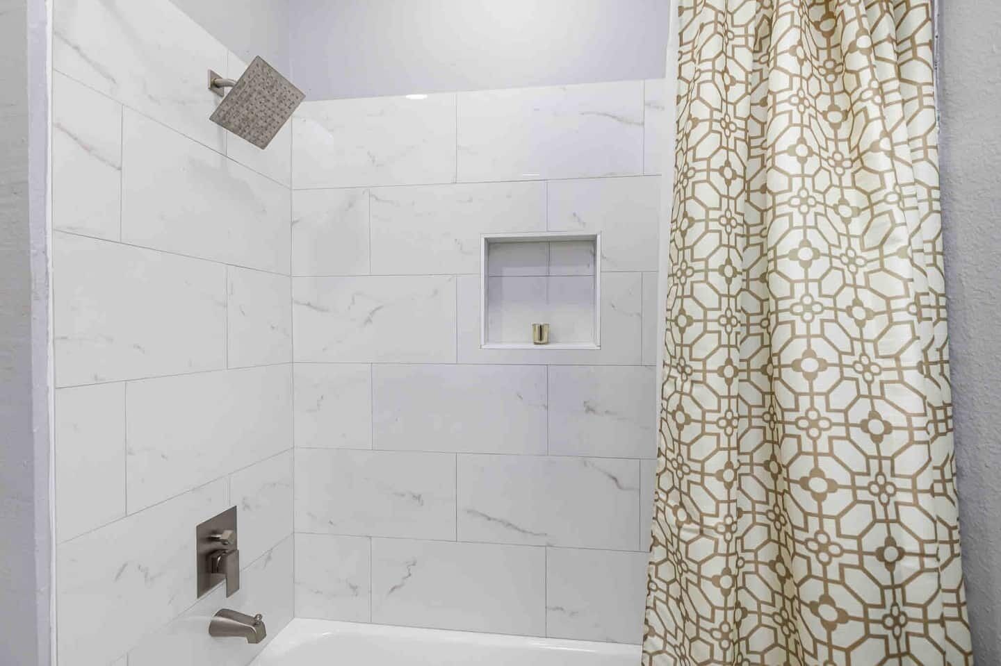 A bathroom with white marble tiles, a shower with a square showerhead, a small shelf, and a patterned shower curtain.