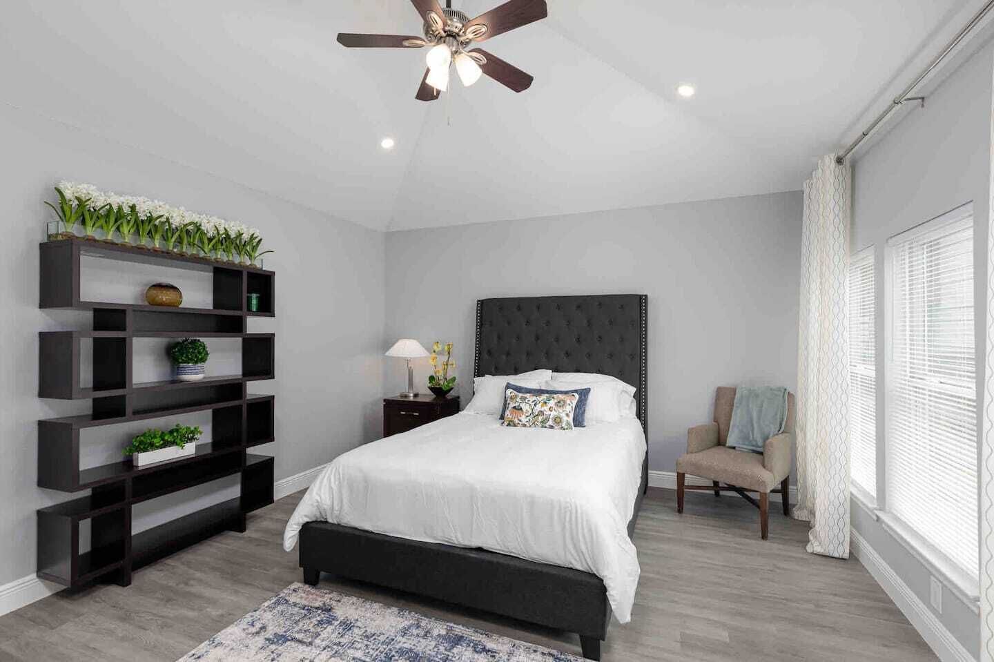 Une chambre moderne avec un grand lit, un ventilateur de plafond, des étagères intégrées ornées de plantes, une table de chevet avec une lampe et une chaise près de la fenêtre.