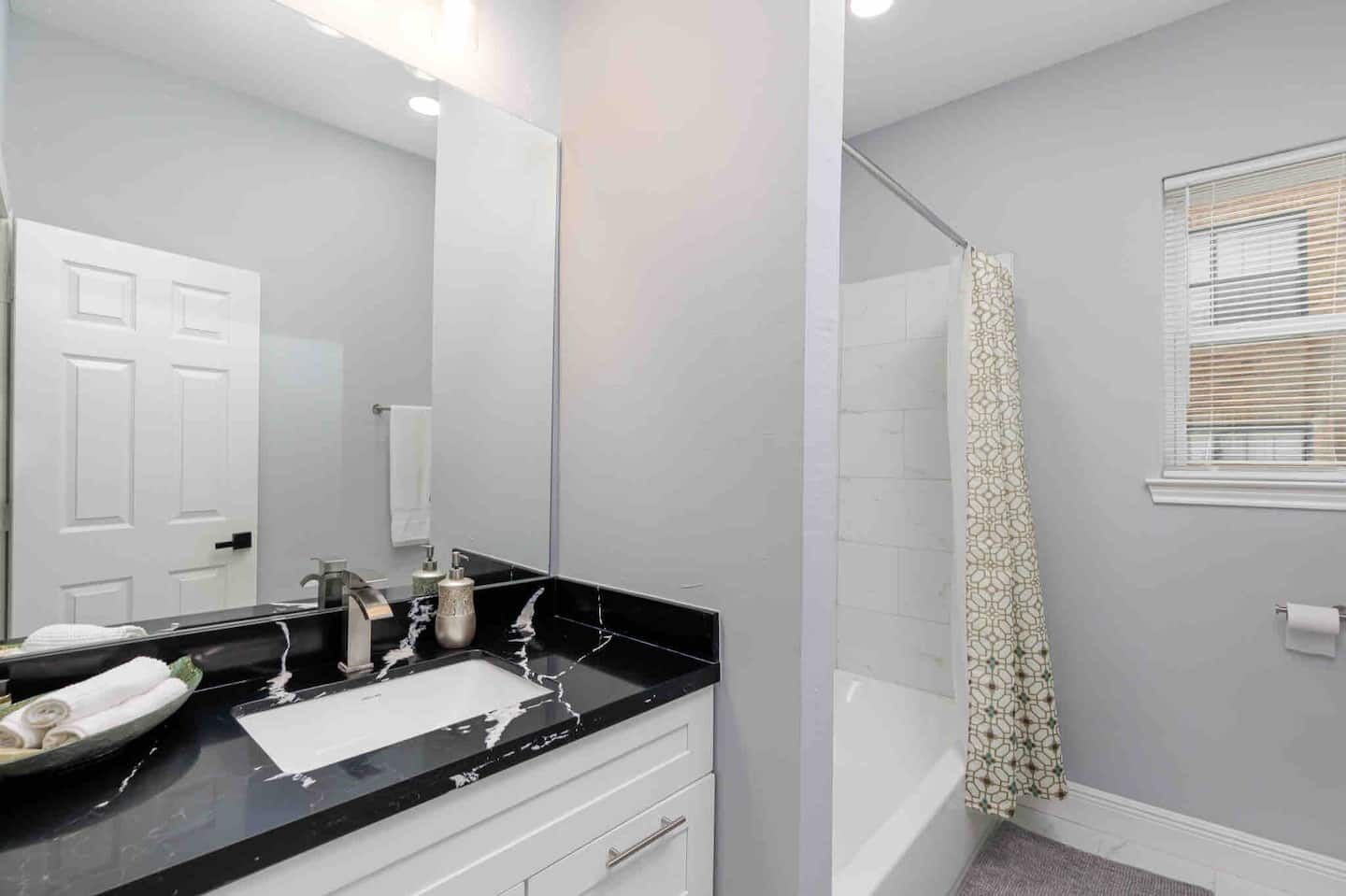 A bathroom with a black countertop, white walls, a white door, a shower with a patterned curtain, and a window with blinds.