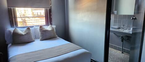 Une chambre d’hôtel avec un lit, deux oreillers, une fenêtre et une salle de bain équipée d’un lavabo et d’un miroir.