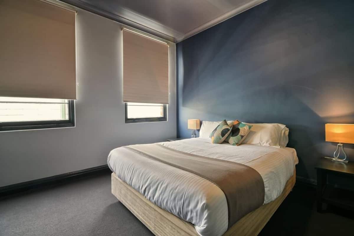 A bedroom with a large bed, two bedside tables with lamps, and two windows with blinds.