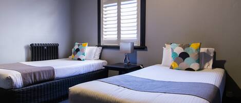 A hotel room with two beds, a nightstand, a lamp, and a window with blinds.