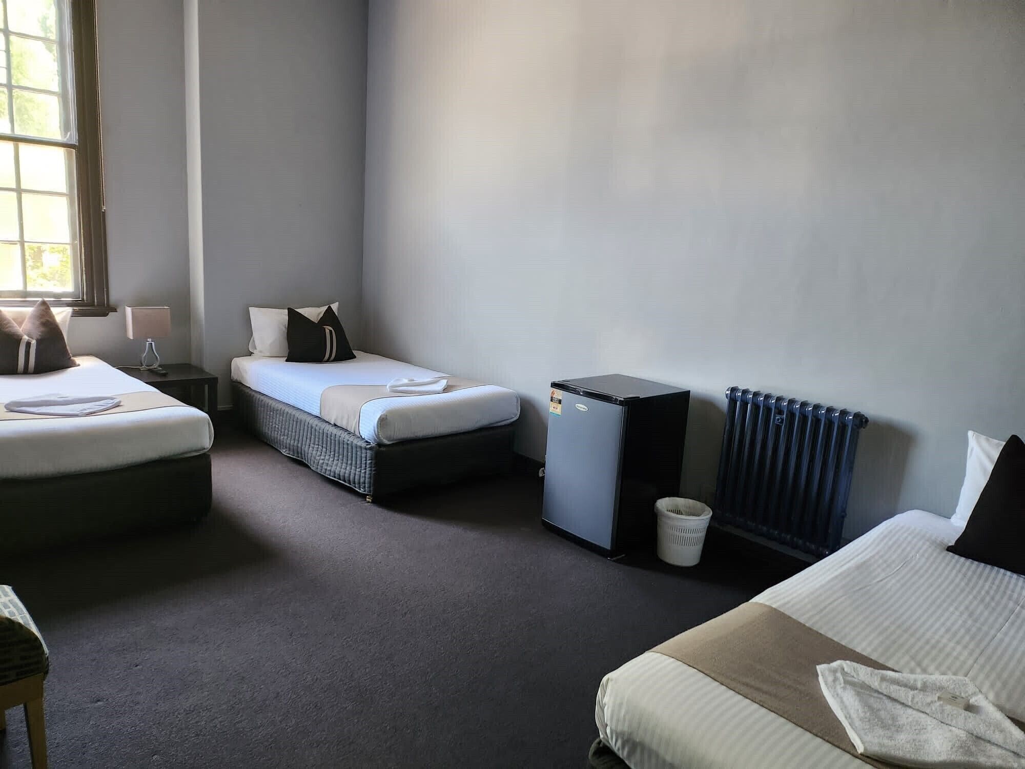 Quarto de hotel com duas camas, um pequeno frigorífico, um radiador e uma janela com luz natural.