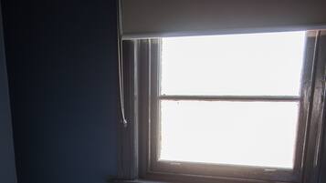 A bed with white bedding and a colorful pillow, positioned under a window with blinds.