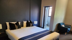 A large bed with a dark headboard, a bedside table with a lamp, a chair, and a view of a bathroom through an open door. - Hotel Canobolas (Orange)