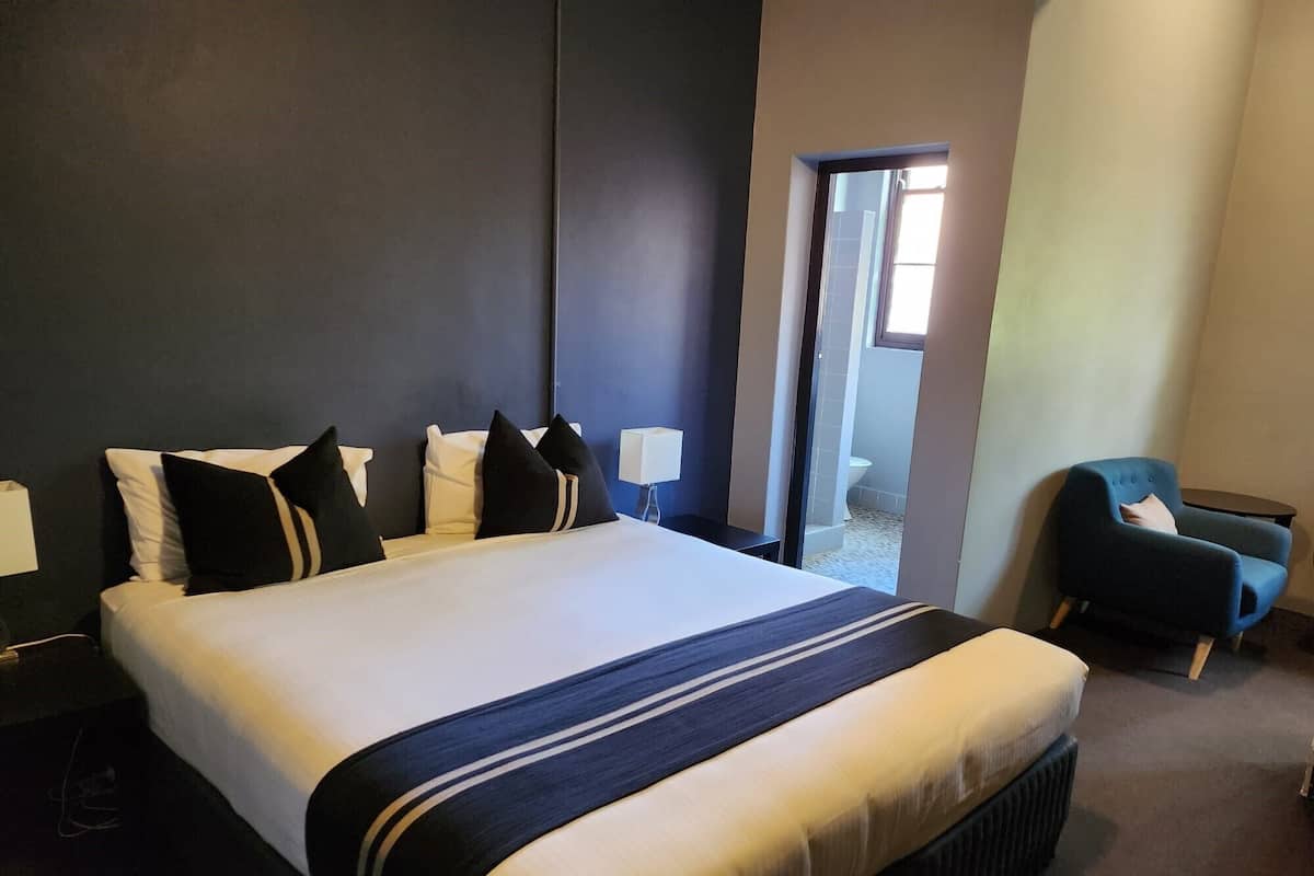 A large bed with a dark headboard, a bedside table with a lamp, a chair, and a view of a bathroom through an open door.