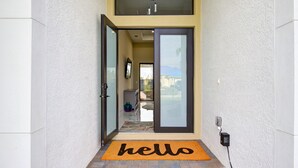 A modern entrance with a sliding glass door, a doormat with "hello" text, and a visible outdoor area with a patio and a mountain view. - Sea Turtle Retreat - Luxurious 4 Bedroom/2 Bath, Electric Heated Pool with Fenced in Yard (Cape Coral)
