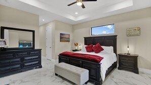 A bedroom with a large bed, a nightstand, a dresser, a bench, a ceiling fan, a TV, and a painting. - Sea Turtle Retreat - Luxurious 4 Bedroom/2 Bath, Electric Heated Pool with Fenced in Yard (Cape Coral)