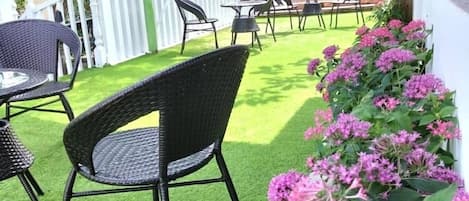 Un espace extérieur aménagé en terrasse, avec un revêtement de gazon synthétique, des meubles en osier et des plantes à fleurs roses.