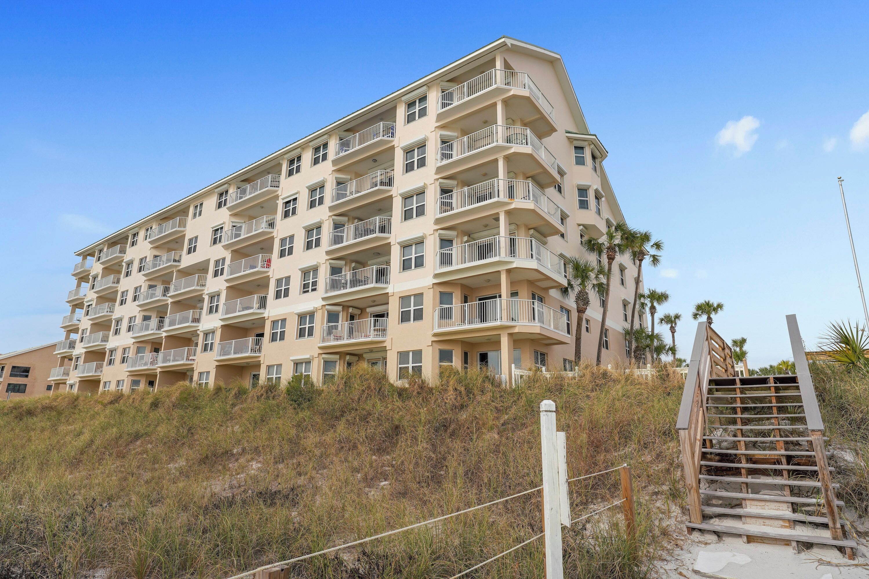 A multi-story hotel with balconies, located near a beach with a wooden staircase leading up to it.