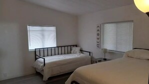 A bedroom with two single beds, a small bedside table, a wall-mounted lamp, and a window with blinds. - The Green River House (Bullhead City)