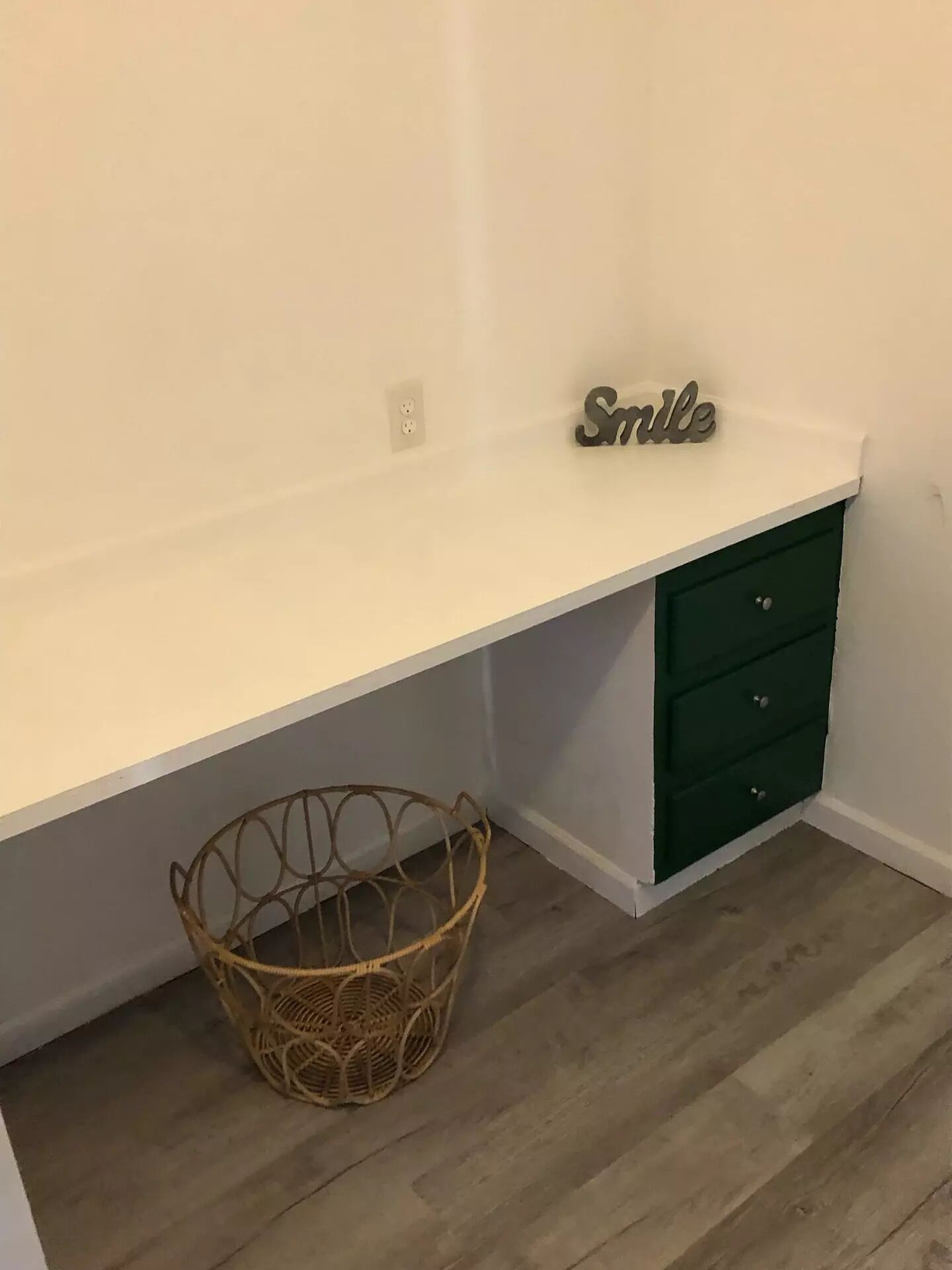 A white desk with a green drawer unit, a wicker basket, and a 'Smile' sign on the desk.