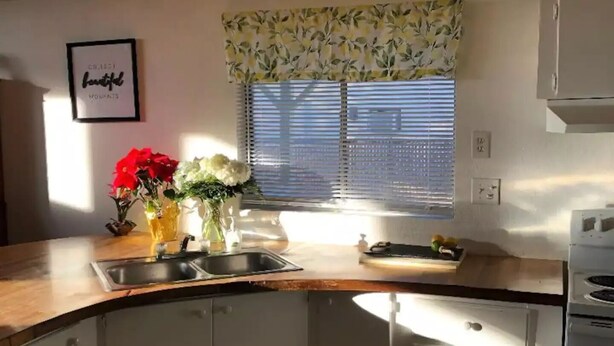 A kitchen with white cabinets, a wooden countertop, a sink, and a window with blinds. - The Green River House (Bullhead City)