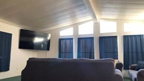 A living room with a large flat-screen TV mounted on the wall, a dark-colored sofa, and a smaller couch. There are windows with curtains, allowing natural light into the room. - The Green River House (Bullhead City)