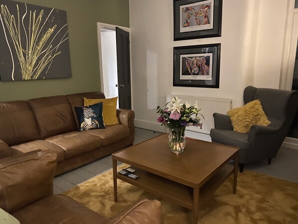 A living room with a brown leather sofa, a wooden coffee table, and a grey armchair. There are framed pictures on the wall and a vase with flowers on the coffee table.