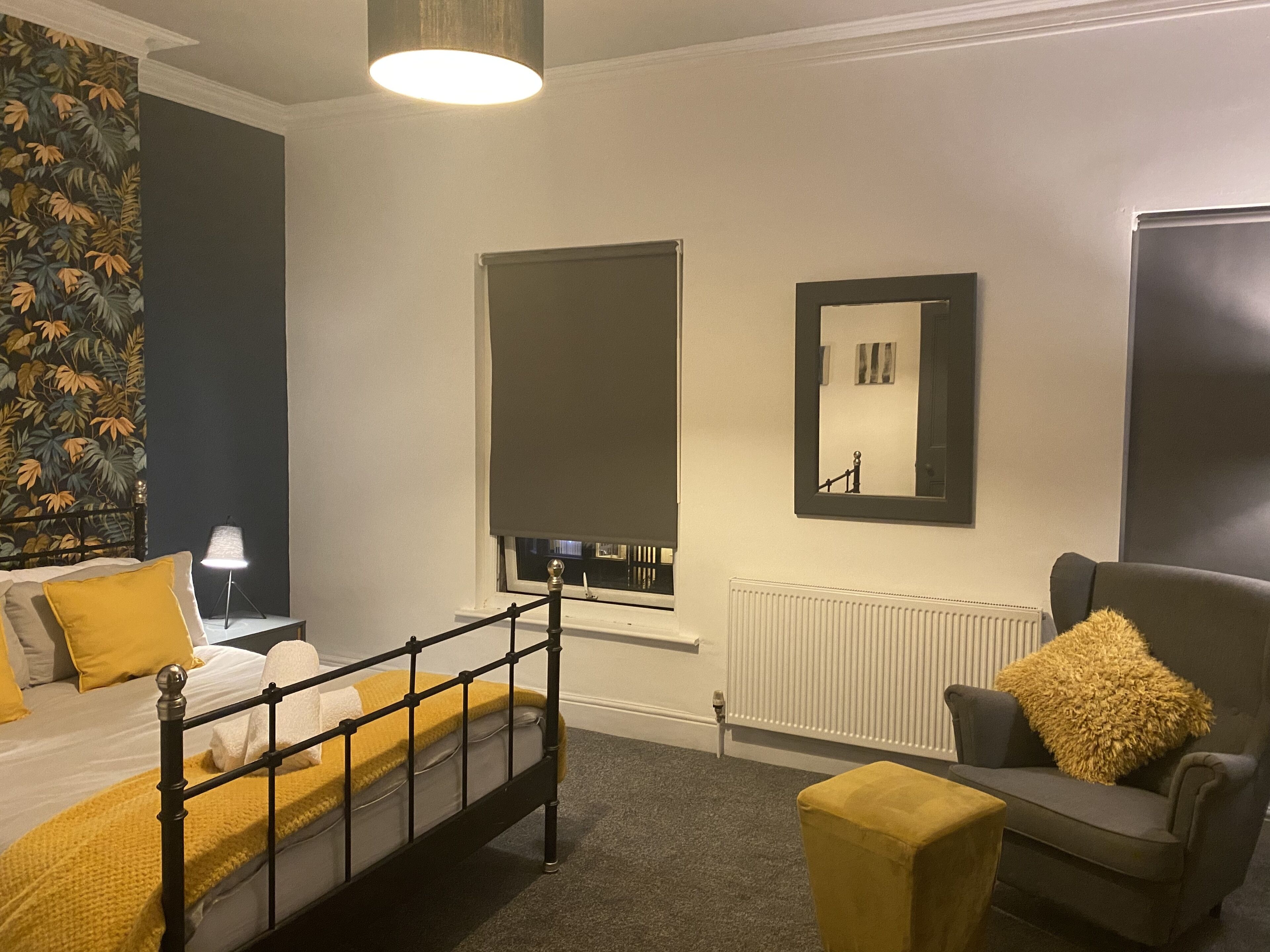 A bedroom with a bed, a nightstand, a lamp, a chair, a sofa, a window with a view, a radiator, and a mirror on the wall.
