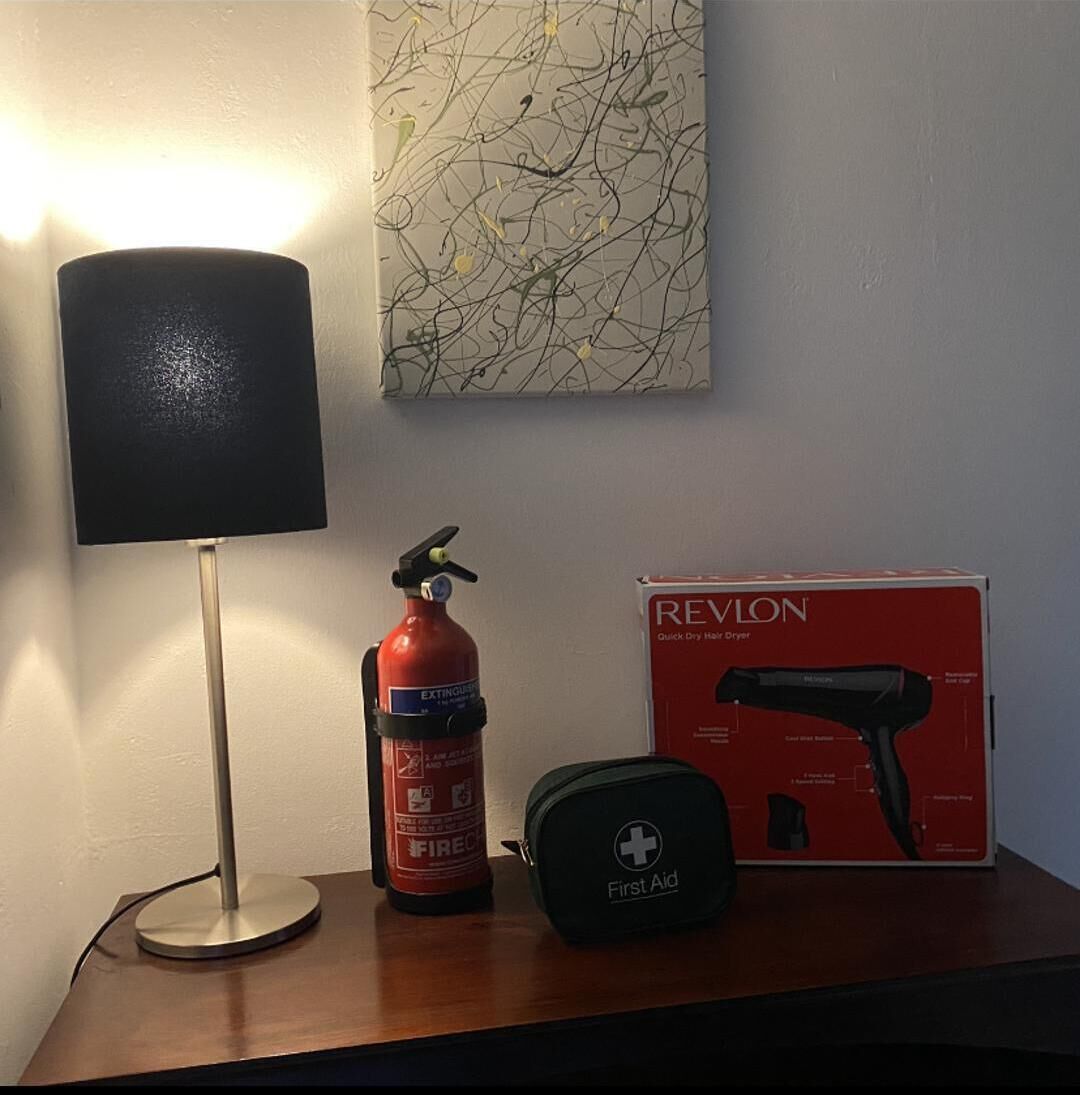 A bedside table with a lamp, a fire extinguisher, a first aid kit, and a Revlon hair dryer box.