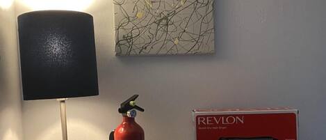 A bedside table with a lamp, a fire extinguisher, a first aid kit, and a Revlon hair dryer box.