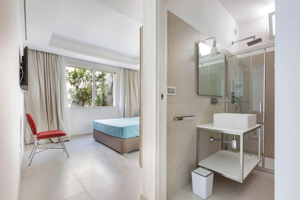 Ein Hotelzimmer mit Bett, Stuhl, Badezimmer mit Waschbecken und Spiegel sowie einem Fenster mit Vorhängen.