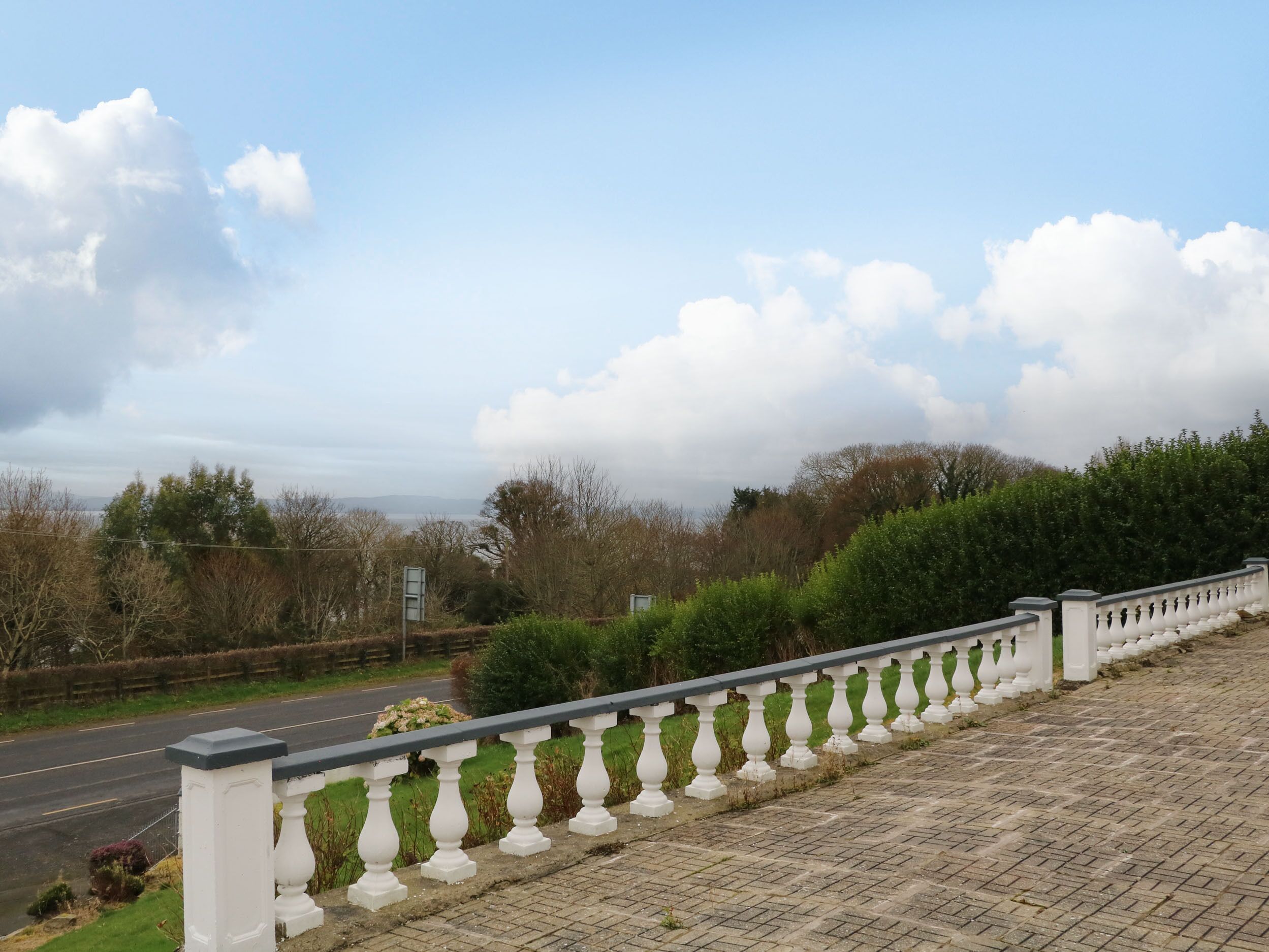 A paved walkway with a white balustrade, a road in the background, and a view of a body of water and distant hills.