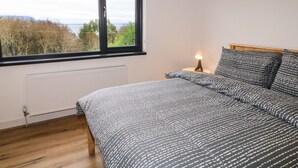 Et soveværelse med et stort vindue, en seng med mønstret sengetøj, et natbord med en lampe og en radiator.