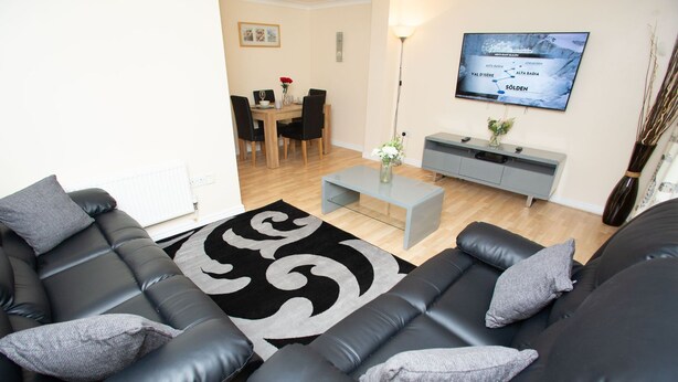 A modern living room with a black leather sofa, a glass coffee table, and a grey console table. There is a flat-screen TV mounted on the wall, a dining area with a wooden table and chairs, and a vase with flowers on the console table.