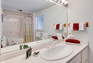 Un baño con un lavamanos blanco, un espejo y una cortina de ducha. Hay toallas colgadas en un perchero y una ventana con un diseño decorativo.