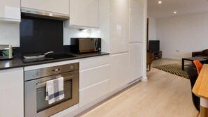 A modern kitchen with white cabinets, a built-in oven, a microwave, and a stove. There is a dining area with a table and chairs, and a television in the background. - 2-bed Apartment Only 15 Mins From Central London (Wembley)