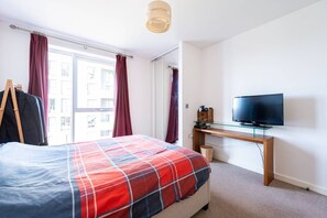 Une chambre à coucher avec un lit, un bureau avec une télévision et une fenêtre avec des rideaux.