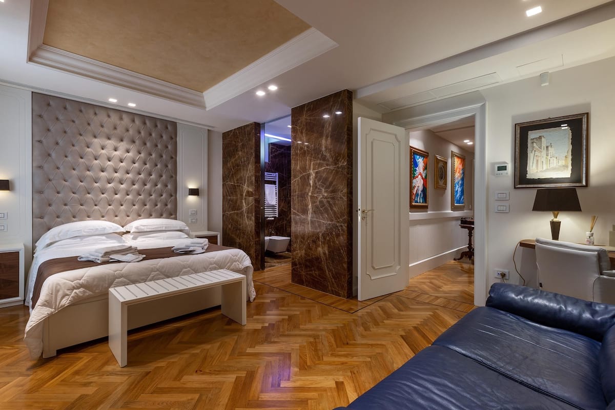 Une chambre moderne avec une tête de lit capitonnée, un parquet en bois disposé en chevrons, un lit avec une literie blanche, un banc, un canapé en cuir bleu et une salle de bain visible à travers une porte ouverte.