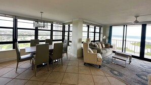 Amplia sala de estar con mesa y sillas de comedor, un sofá y vista al océano a través de grandes ventanales.
