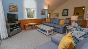 Een woonkamer met een grijze bank, een televisie op een dressoir, een salontafel en een raam met gordijnen.