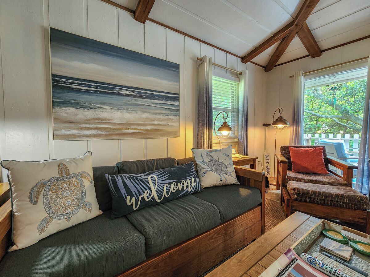 Un salon chaleureux avec un canapé vert, une table basse en bois et une chaise en bois. Un tableau représentant un paysage marin est accroché au mur, ainsi qu’une lampe et une fenêtre avec des rideaux.