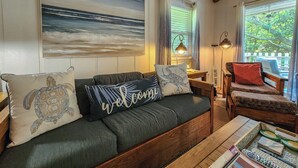 Una sala de estar acogedora con un sofá verde, una mesa de centro de madera y una silla de madera. Hay un cuadro de un paisaje marino en la pared, una lámpara y una ventana con cortinas.