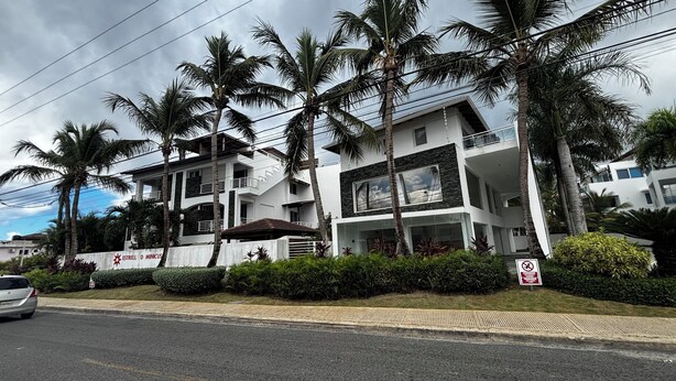 A modern multi-story building with balconies, surrounded by palm trees and greenery.