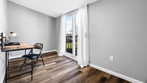 Une pièce avec un plancher en bois, un bureau avec une chaise, une fenêtre avec des rideaux blancs et un luminaire au plafond.