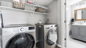 Une buanderie équipée de deux machines à laver, d’une étagère où sont pliées les serviettes, et une salle de bain avec un miroir et un lavabo.