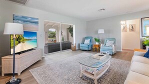 Ein modernes Wohnzimmer mit Fernseher, zwei blauen Sessel, einem Glastisch und einem großen Fenster mit Blick auf einen Pool und Grünflächen.