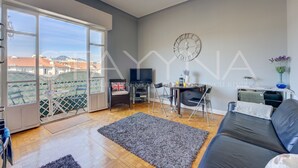 Un salon moderne avec une grande fenêtre, un canapé en cuir noir, un tapis gris, une table à manger avec des chaises, un téléviseur et une horloge murale.