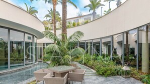 A modern outdoor patio with a glass table and chairs, surrounded by palm trees and a swimming pool. - Spa Du Cap 2 bedroom apartment (Antibes)