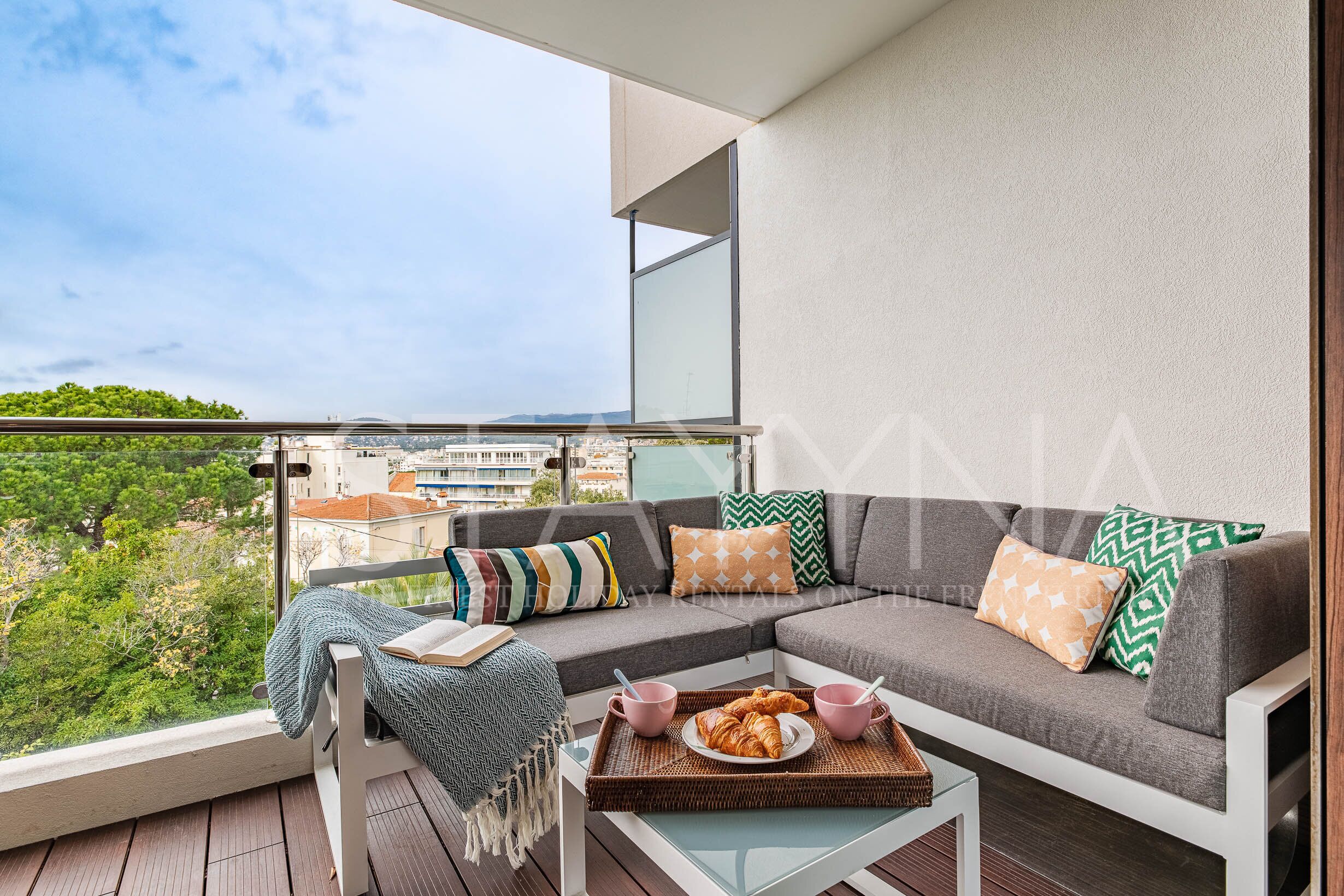 Een balkon met een bank, kussens, een dienblad met croissants en uitzicht op groen en gebouwen.