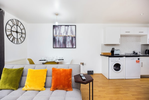 A modern living room with a sofa, a remote control, and a washing machine in the background.