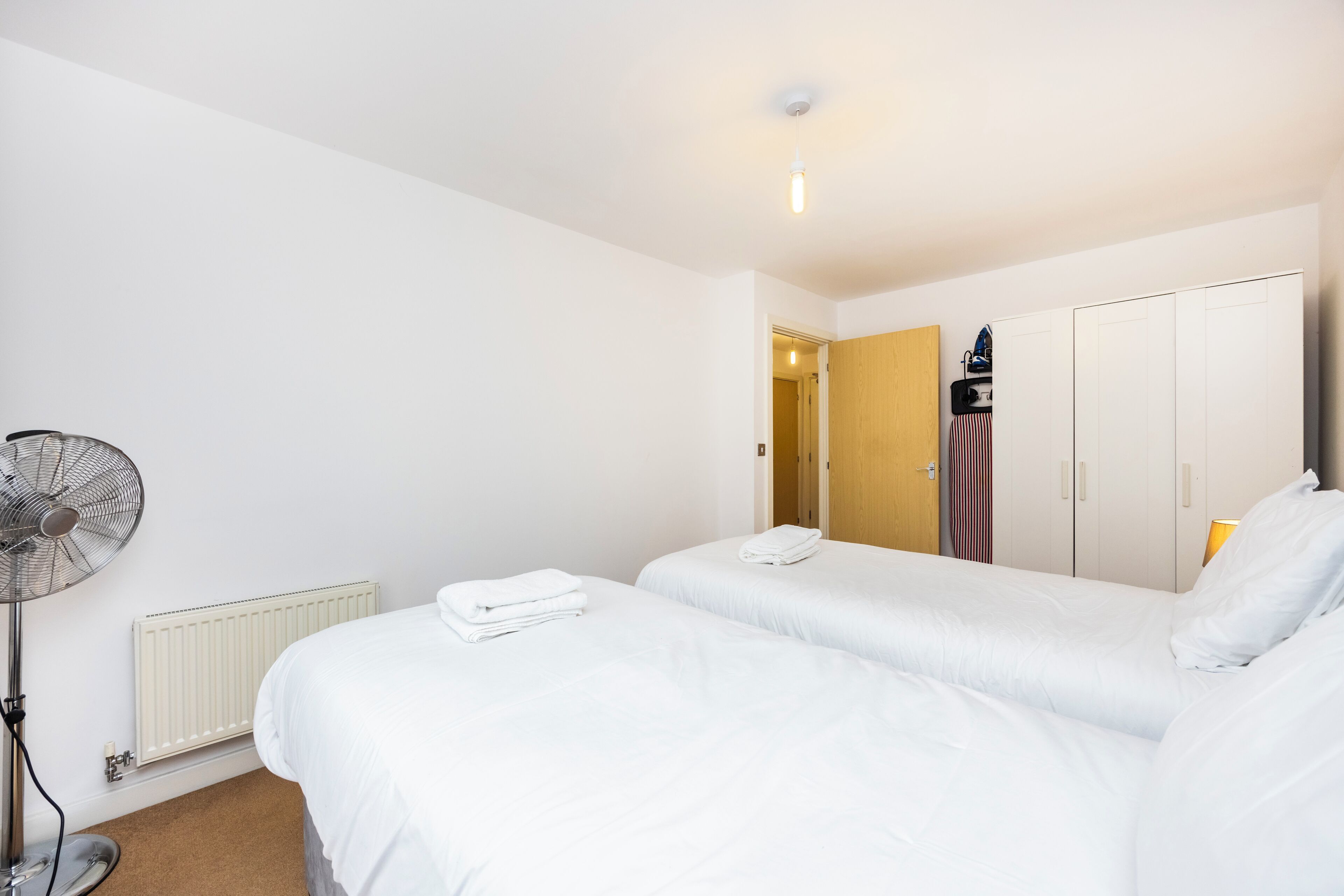 A hotel room with two beds, a radiator, a standing fan, and a wardrobe.