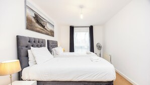 Une chambre avec un grand lit, une table de chevet avec une lampe, une œuvre d’art accrochée au mur, une fenêtre avec des rideaux et un radiateur.