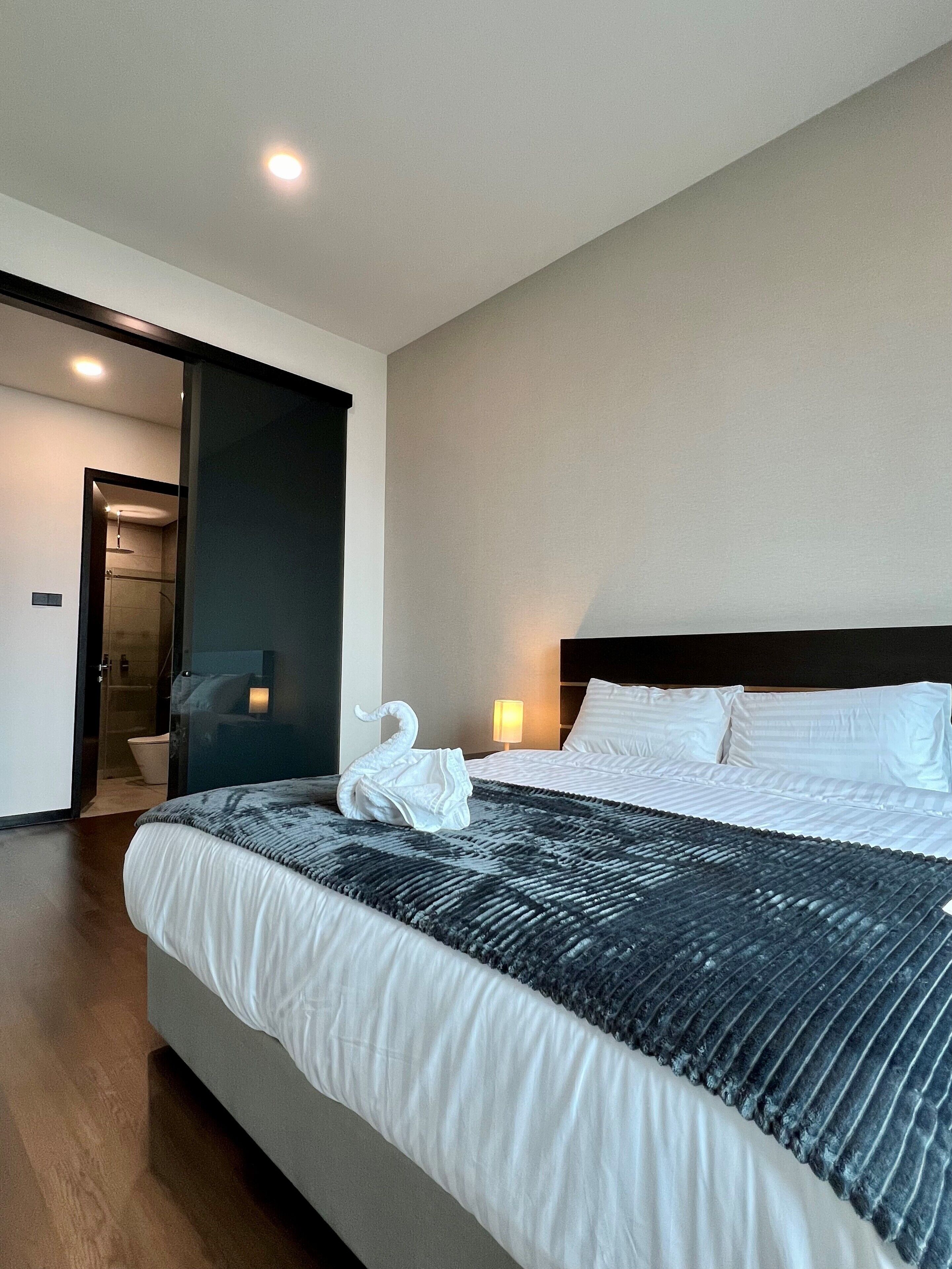 A modern hotel room with a large bed, a dark headboard, a glass partition, a small bathroom, and a nightstand with a lamp.