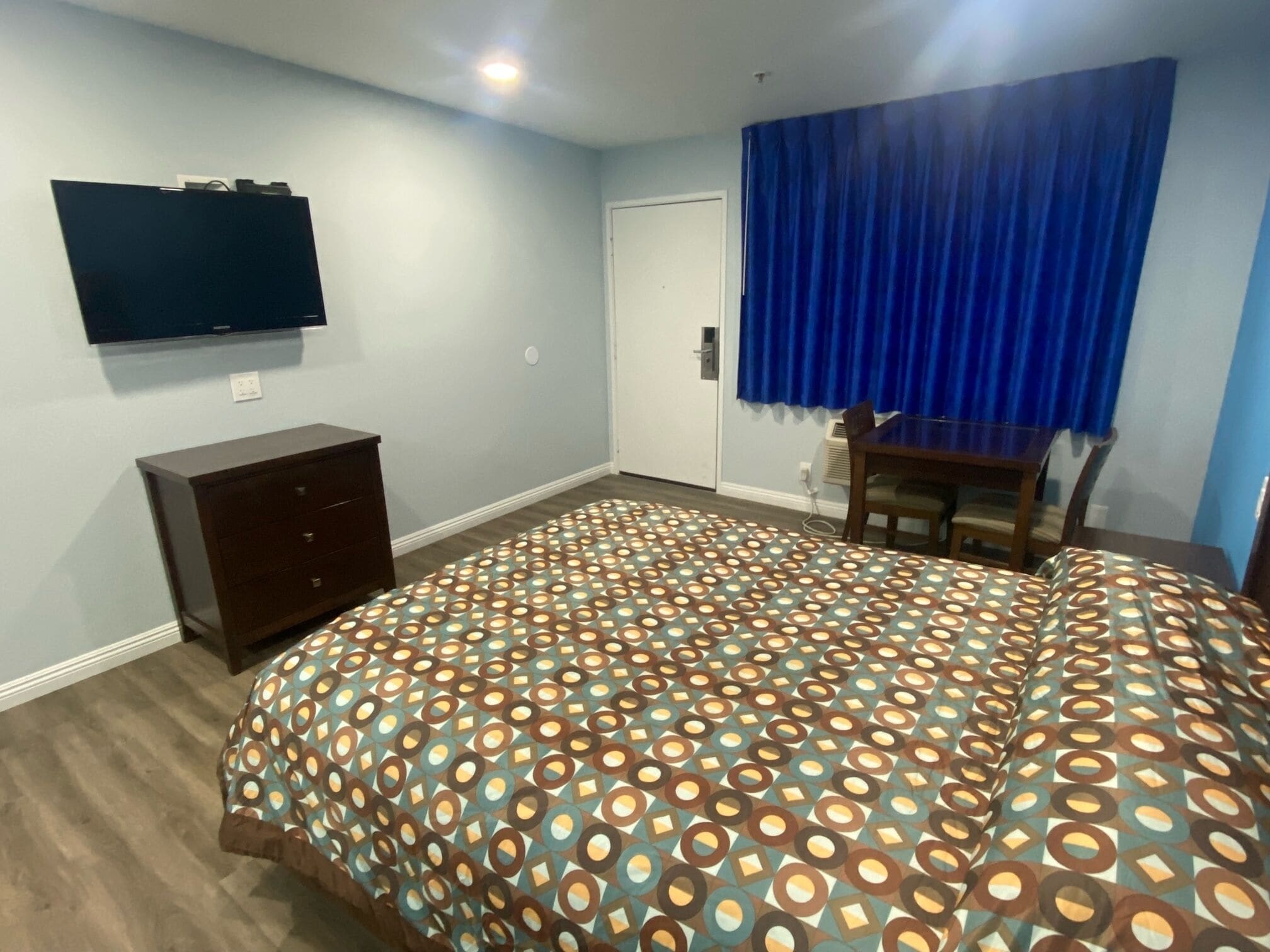 Een slaapkamer met een bed met een geborduurd sprei, een donker houten dressoir, een flatscreen-televisie aan de muur en een raam met blauwe gordijnen.