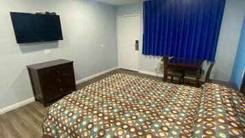 A bedroom with a patterned bedspread, a dark wooden dresser, a flat-screen TV mounted on the wall, and a window with blue curtains.
