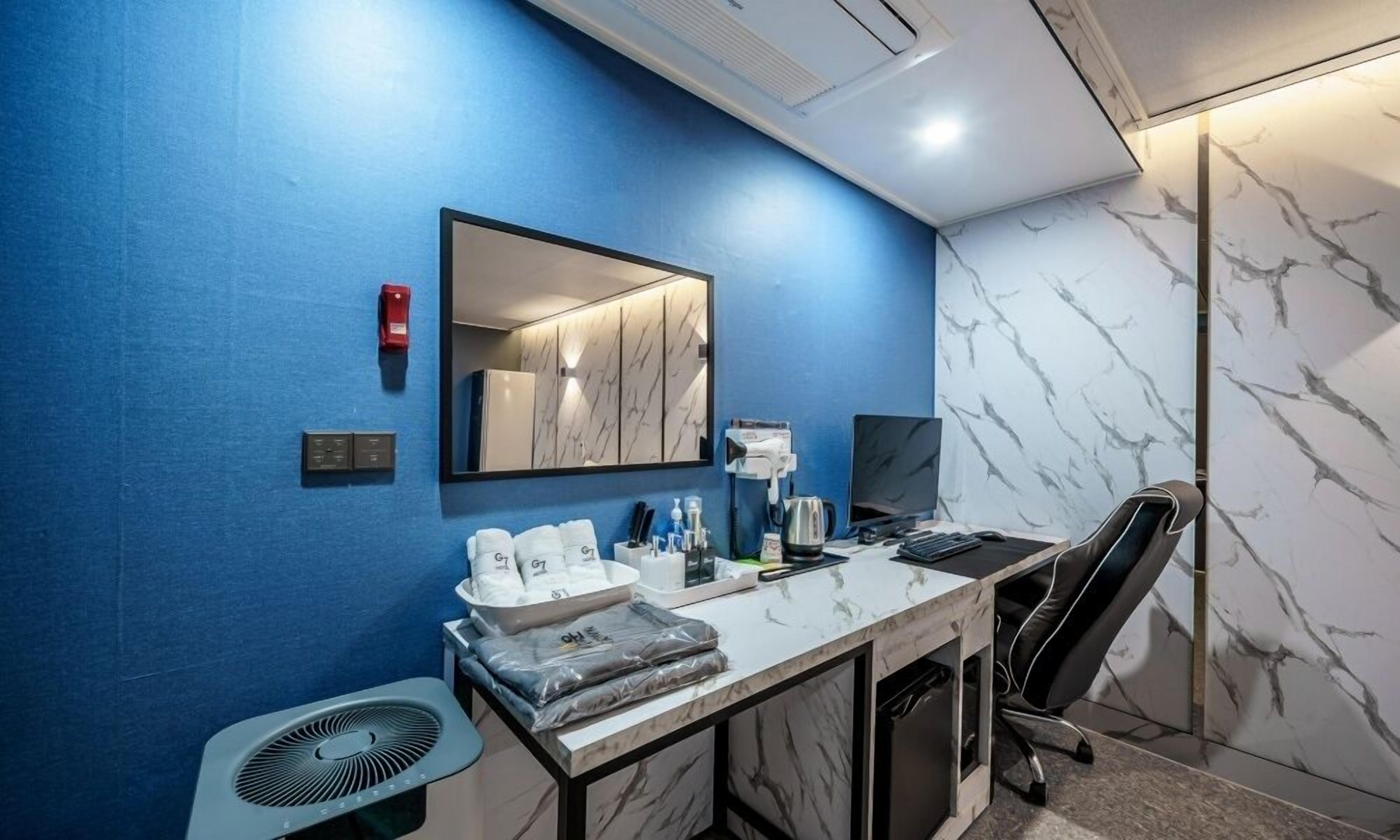 A modern hotel room with a blue accent wall, a marble-patterned wall, a desk with a computer, a chair, a fan, and a mirror.